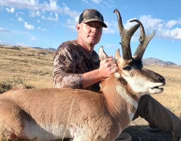 A hunter proudly poses with a pronghorn antelope, its distinctive black horns curved backward, in the open Wyoming plains under a bright blue sky.